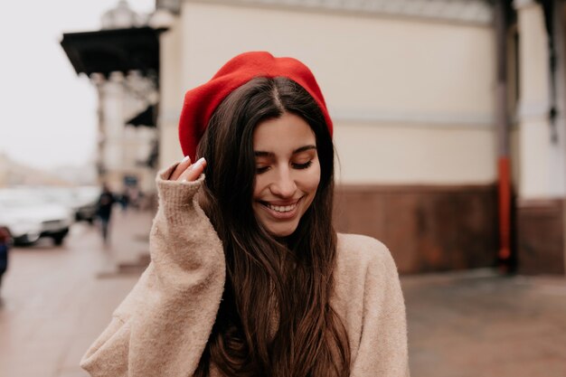 Романтичная застенчивая женщина с длинными темными волосами в красной кепке и бежевой куртке идет по улице