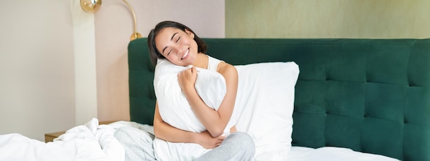 Бесплатное фото Романтическая улыбающаяся азиатская девушка лежит в постели, обнимает подушку и смотрит мечтательным ленивым утром в