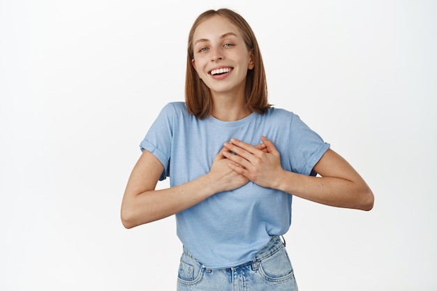 Romantic smiling blond woman holding hands on heart, smiling passionate and gazing at camera, fall in love, thrilled and delighted with smth, standing against white background.