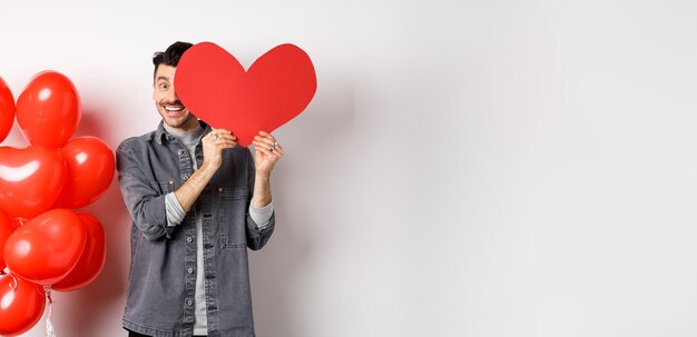 無料写真 ロマンチックな笑顔の男はバレンタイン ハート カードと顔を覆い、lo を祝うカメラで幸せそうに見えます
