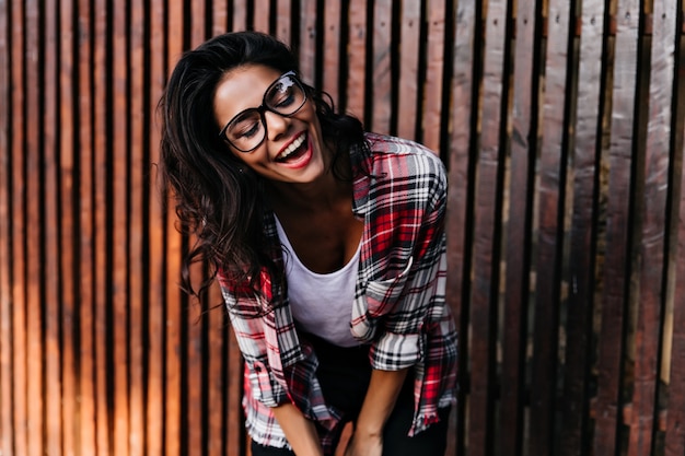 Romantic tanned girl laughing while posing near wooden fence. Happy dark-haired lady in checkered shirt smiling with eyes closed.