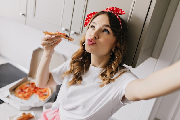 Бесплатное фото Романтичная белая девушка делает селфи во время еды пиццы. крытый снимок кудрявой кавказской дамы дурачиться во время завтрака.