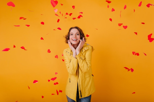 Romantic white girl with cute face expression posing with red hearts. Indoor photo of curly young woman celebrating valentine's day with smile.