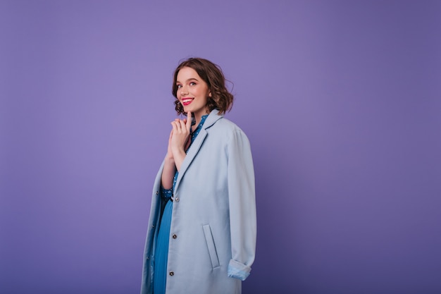 Romantic white woman in elegant coat playfully posing on purple wall. Indoor photo of joyful curly female model with short haircut.