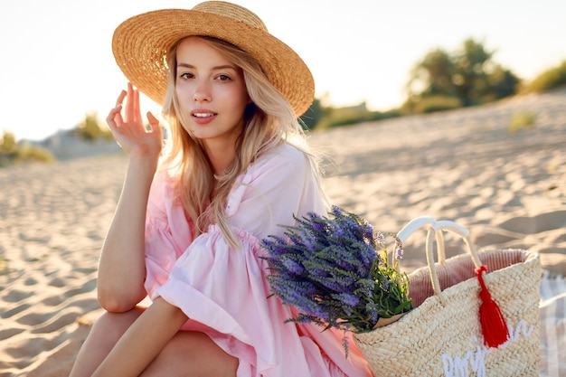 トレンディな帽子とビーチでポーズをとるエレガントなピンクのドレスのロマンチックな白人女性。ストローバッグと花の花束を保持しています。