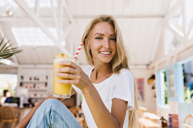 Romantic woman in blue jeans drinking orange cocktail with pleasure. Indoor shot of smiling blonde girl holding glass of cold juice while sitting in cafeteria.
