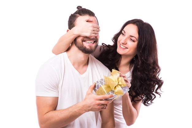 Romantic woman covering her boyfriend's eyes. Woman standing behind man with gift isolated on grey background