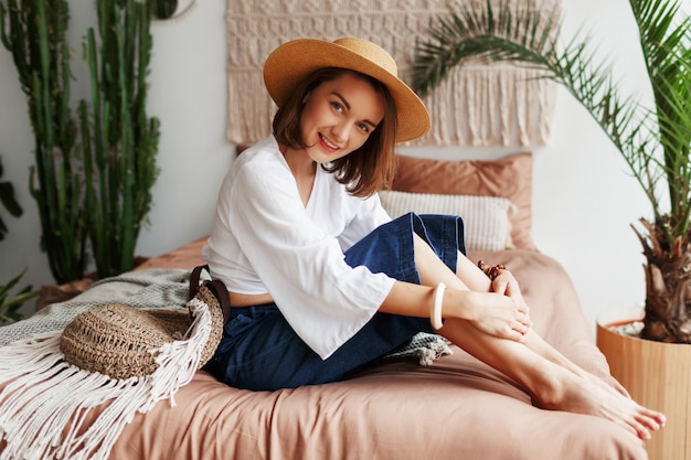 Romantic woman with candid smile sitting on bed, enjoying sunny morning in her stylish flat in boho style