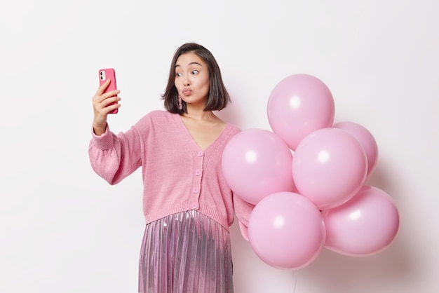 Romantic young Asian woman keeps lips rounded poses for making selfie enjoys holiday holds bunch of helium balloons dressed in festive clothes isolated over white background prepares for party