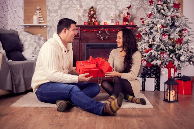 クリスマスの日に暖炉の前に座っているロマンチックな若いカップル。クリスマスプレゼント。伝統的なクリスマスの飾り。