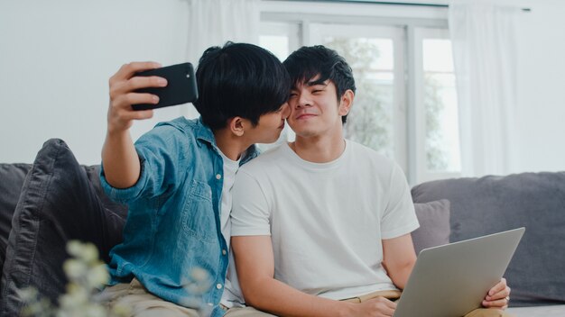 Romantic young gay couple funny selfie by cellphone at home. Asian lover LGBT male happy relax fun using technology mobile phone smiling take a photo together while lying sofa in living room .