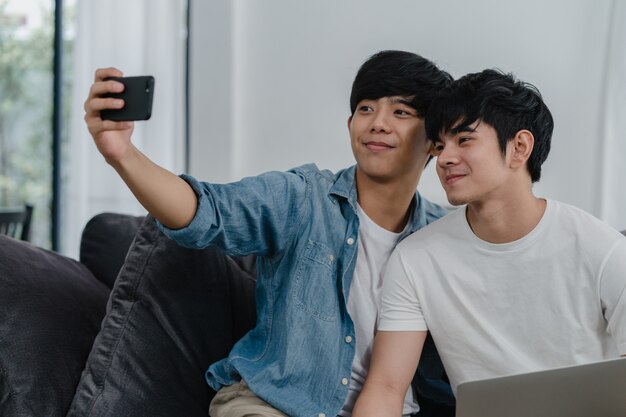 自宅で携帯電話でロマンチックな若い同性愛者カップル面白いselfie。アジアの恋人男性の幸せは、リビングルームでソファに横たわっている間一緒に写真を撮る笑顔技術携帯電話を使用して楽しいリラックスします。
