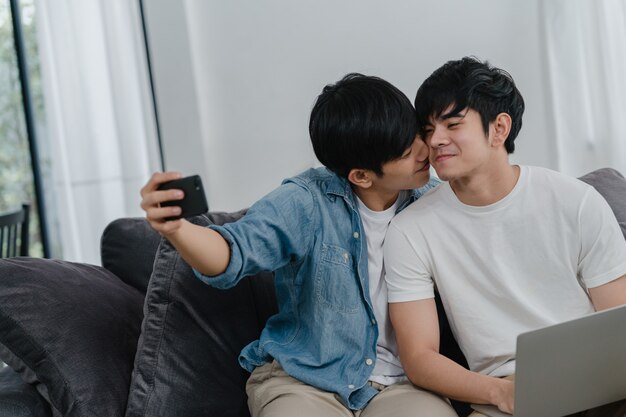 Romantic young gay couple funny selfie by cellphone at home. Asian lover male happy relax fun using technology mobile phone smiling take a photo together while lying sofa in living room .