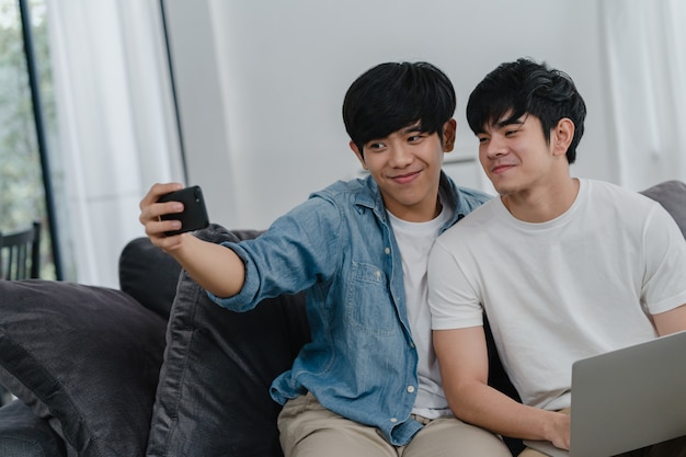 Romantic young gay couple funny selfie by cellphone at home. Asian lover male happy relax fun using technology mobile phone smiling take a photo together while lying sofa in living room .