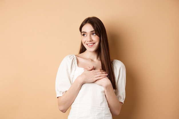 Romantic young girl in dress holding hands on heart, smiling and looking at empty space thankful, feeling gratitude, standing on beige background