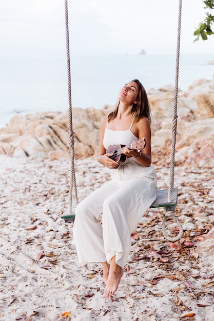 Romantic young happy calm caucasian woman with ukulele on tropical rocky beach at sunset