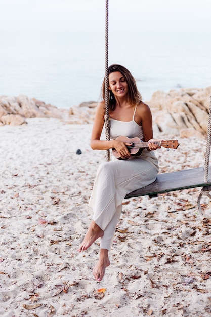 Romantic young happy calm caucasian woman with ukulele on tropical rocky beach at sunset