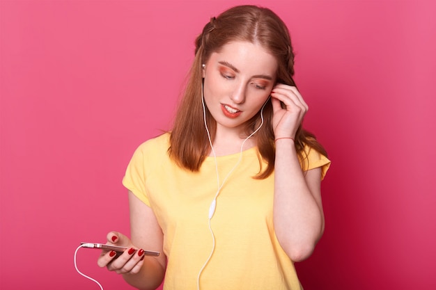 かわいい髪型とプロのロマンチックな若い女性が彼女のイヤホンを右に固定し、片手で軽い携帯電話を持ちます。明るいピンクに分離されたモデルのポーズ。