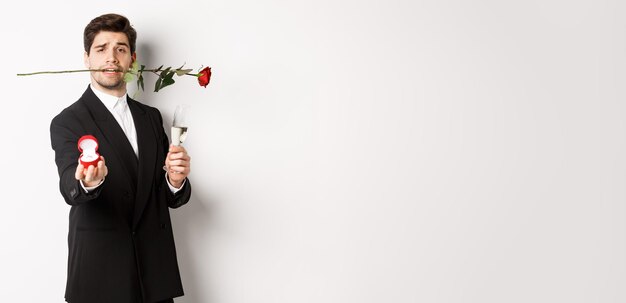 Free photo romantic young man in suit making a proposal holding rose in teeth and glass of champagne showing en