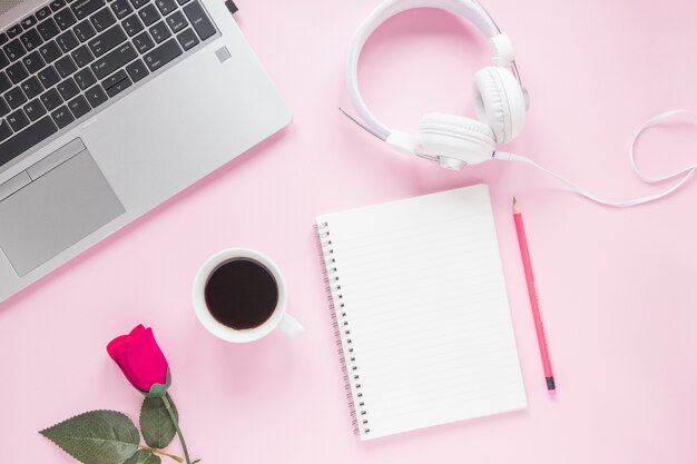 Rose; coffee cup; headphone; laptop; spiral notepad and pencil on pink background