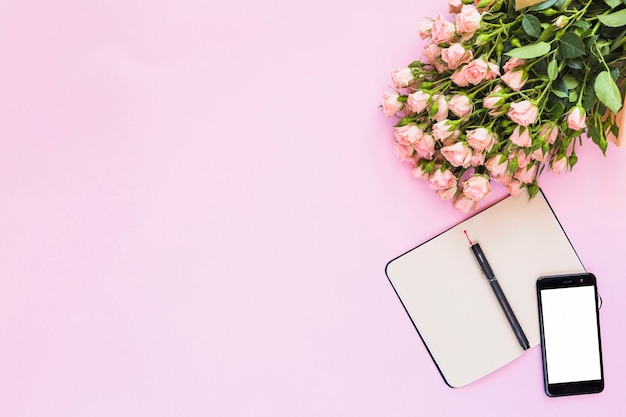 Mazzo delle rose con un diario in bianco aperto con la penna e smartphone su fondo rosa