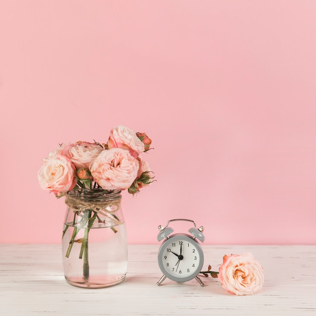 Roses vase near the alarm clock on wooden desk against pink background