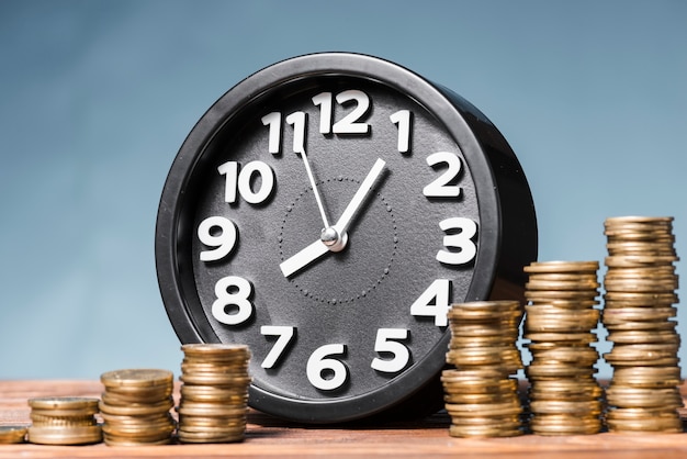 Round alarm clock with stack of increasing coins against blue background