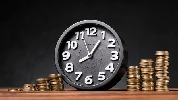 Round clock between the increasing coins on wooden desk against black backdrop