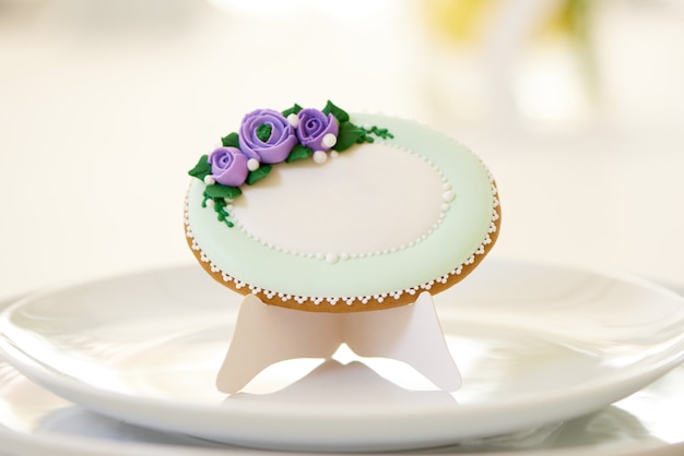 Round gingerbread, covered with white glaze and decorated with violet flowers stands and pattern on the plates,near a wine glasses on a festive wedding table. Photo was made
