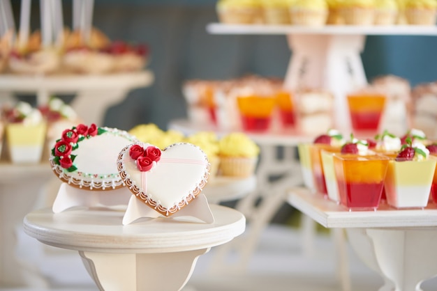 Round and heart shaped glazed cookies, decorated with glaze flowers and pattern are on the wooden stand in the restaurant. There is a colorful delicious candybar behind them. Good choice for wedding.