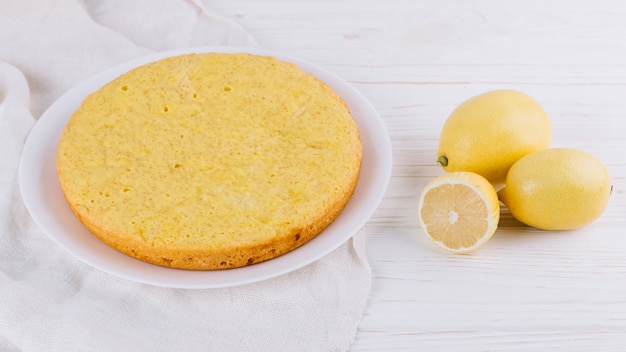 Круглый лимонный пирог подается в белой тарелке с целыми лимонами на деревянном фоне