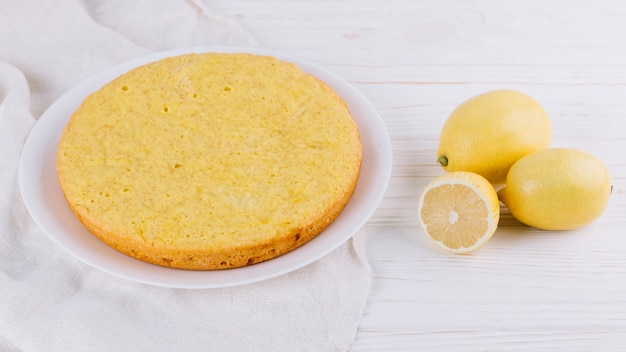 Round lemon cake served in white plate with whole lemons on wooden backdrop