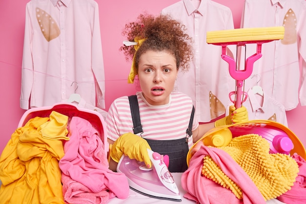 Routine work and household concept. Worried impressed housewife with curly hair holds mop poses near ironing board with electric iron tidies up clothes brings pile of laundry busy doing housework