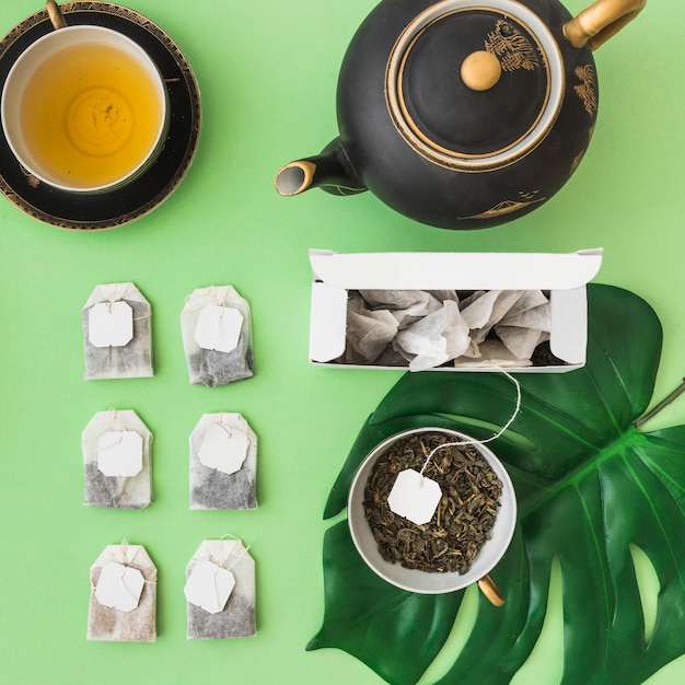 Row of different tea bags with tea cup and teapot on pale green backdrop