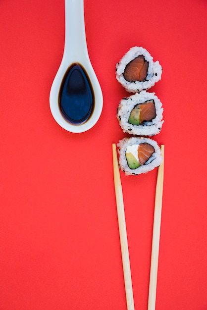Row of sushi rolls with chopsticks and soya sauce in white spoon over red backdrop