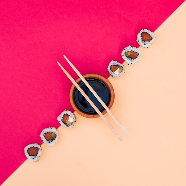 Row of sushi with soya sauce in bowl with chopsticks on pink and yellow dual backdrop