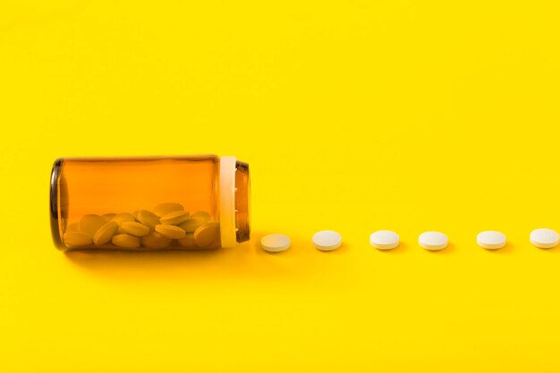 Ряд белых таблеток перед открытой стеклянной бутылкой над желтым фоном