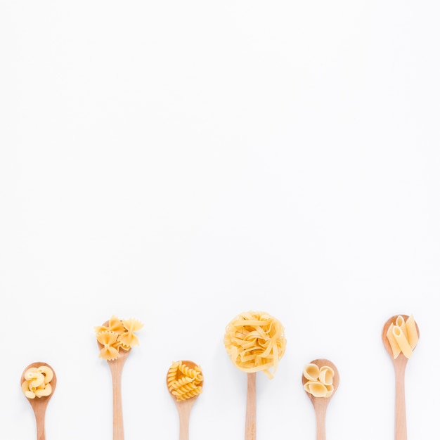 Row of wooden spoon with various dry pasta on white surface