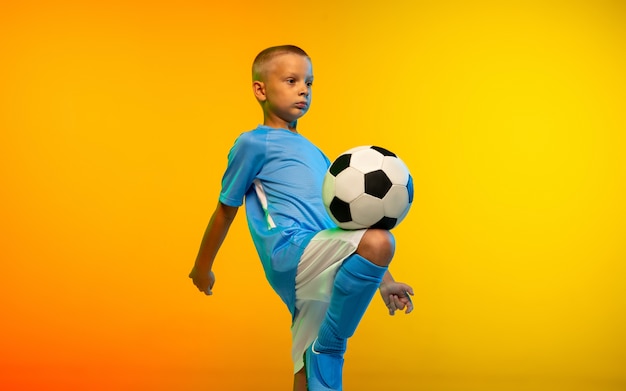 Run. Young boy as a soccer or football player in sportwear practicing on gradient yellow studio