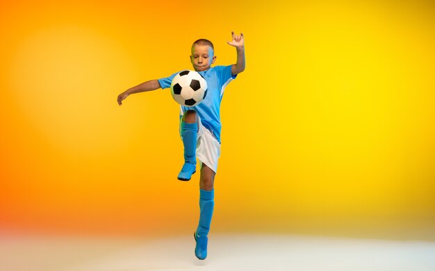 Run. Young boy as a soccer or football player in sportwear practicing on gradient yellow studio