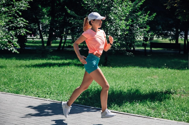 Running smiling young sporty woman in park in sportswear on background of green trees and lawn on gr...