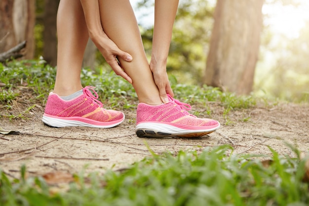 Running sport injury. Female athlete jogger wearing pink sneakers touching her twisted or sprained ankle while jogging or running outdoors.