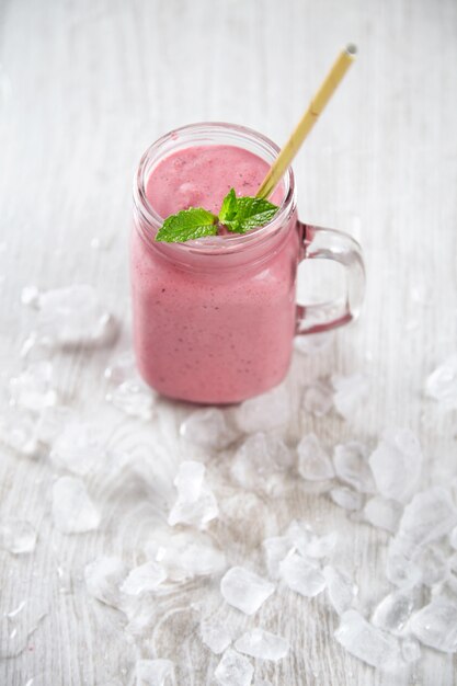 rustic jar with cold fresh berry smoothie isolated on wooden table with golden straw inside surrounded with crashed melted ice cubes