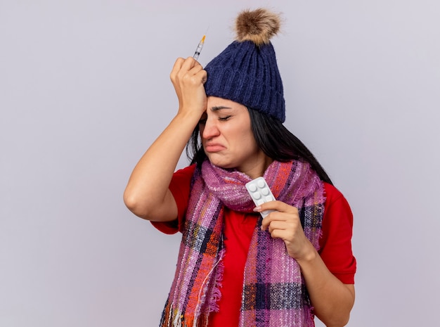 Sad and aching young ill woman wearing winter hat and scarf holding syringe and pack of tablets keeping hand on head isolated on white wall