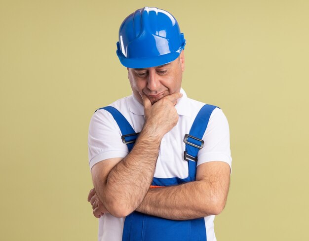 Sad adult builder man in uniform puts hand on chin and looks down isolated on olive green wall