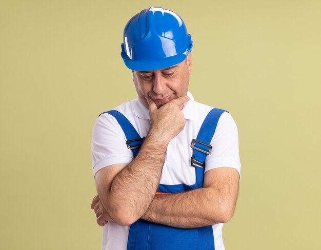 Грустный взрослый строитель в униформе кладет руку на подбородок и смотрит вниз изолированно на оливково-зеленой стене