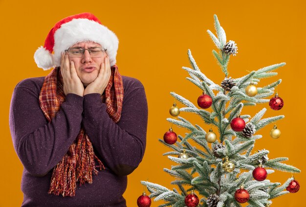 오렌지 배경에 고립 된 측면을보고 얼굴에 손을 유지 장식 된 크리스마스 트리 근처 목 서 스카프와 안경과 산타 모자를 쓰고 슬픈 성인 남자