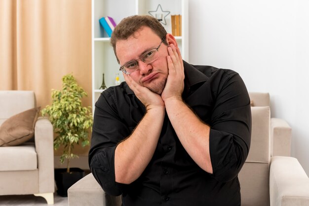 sad adult slavic man in optical glasses sits on armchair putting hands on face