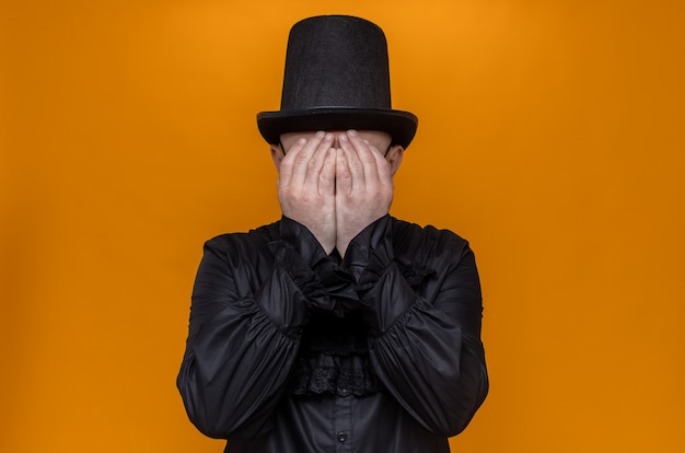 Free photo sad adult slavic man with top hat and optical glasses in black gothic shirt covering his face with hands