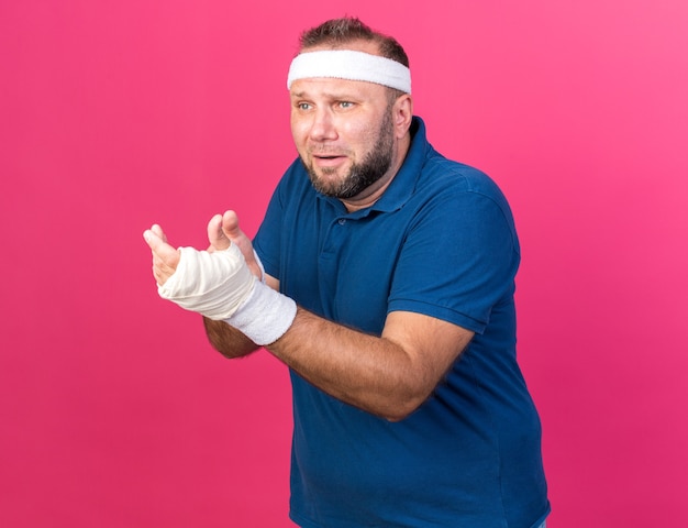 Free photo sad adult slavic sporty man wearing headband and wristbands looking and pointing at side isolated on pink wall with copy space